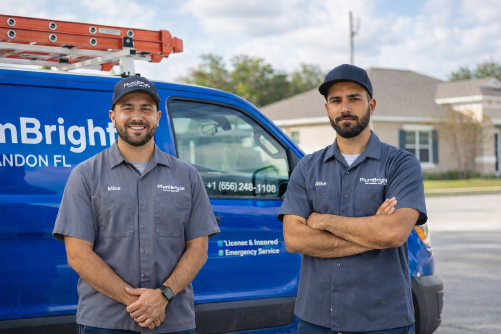 **Alt Text:** Two professional plumbers in grey uniforms standing in front of a blue plumbing service van outside a residential area.